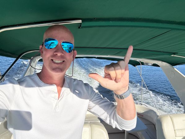 Smiling person in blue mirrored sunglasses and a white henley making a shaka sign aboard a motorboat under a green canopy, wake trailing across open water on a sunny day.