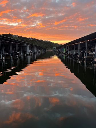 Sunset-splashed sky over a marina with glowing orange clouds reflected like a fiery pathway between covered boat slips on a calm lake.