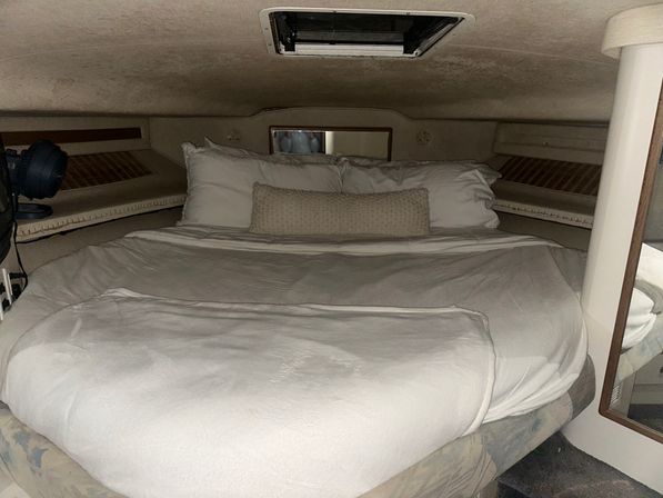 Cozy v-berth boat cabin with white bedding, textured lumbar pillow, overhead hatch and built-in side shelves in a compact marine interior.