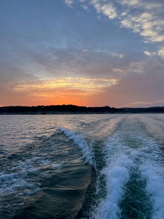 Serene lake sunset at dusk with orange-purple clouds, silhouetted shoreline, and a boat's wake rippling across the water