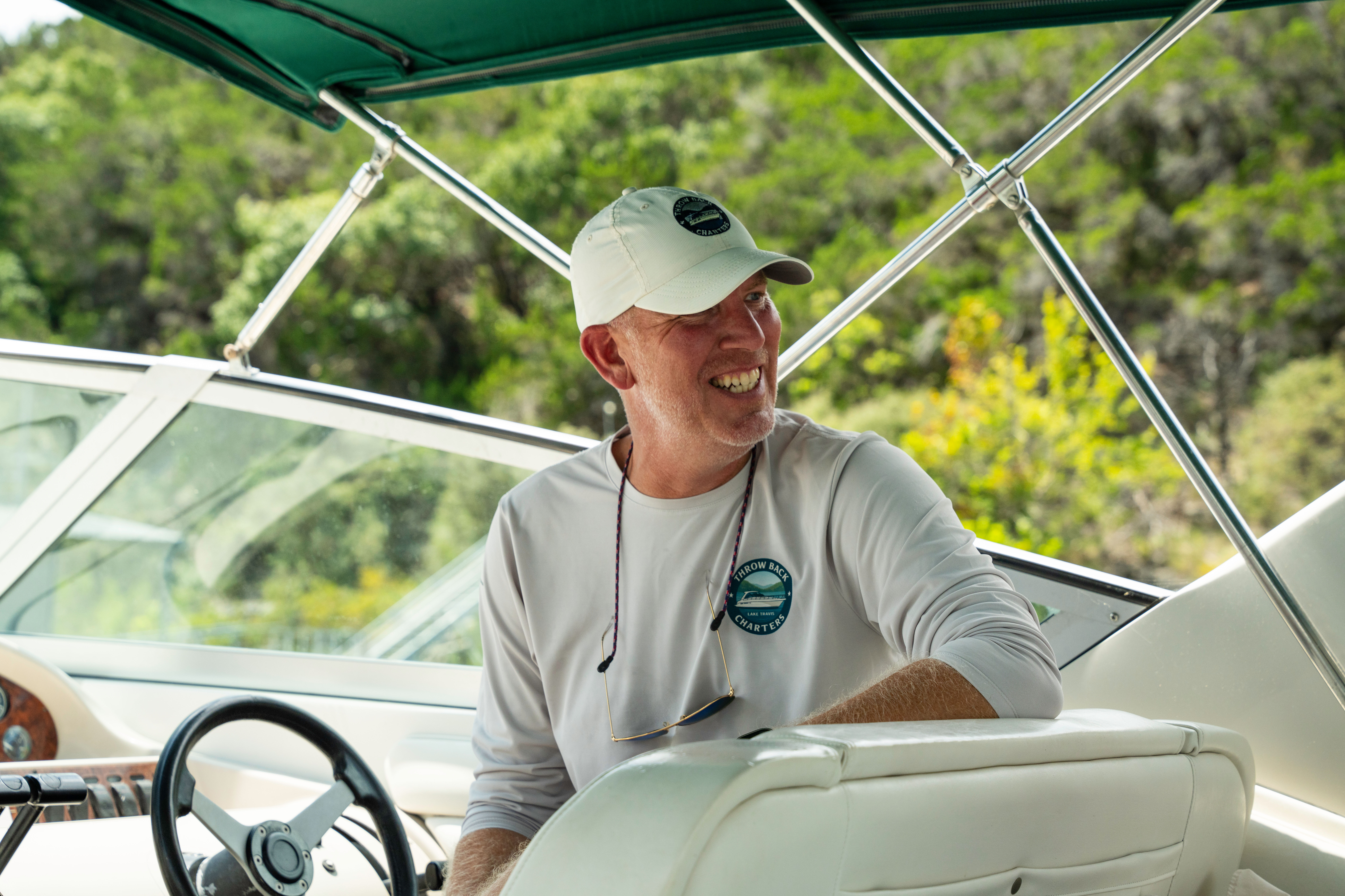 Smiling man at the helm of a motorboat under a green bimini, wearing a white cap and long-sleeve shirt, steering with a forested shoreline in the background