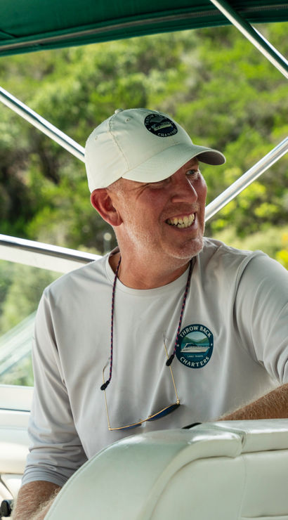 Smiling man at the helm of a motorboat under a green bimini, wearing a white cap and long-sleeve shirt, steering with a forested shoreline in the background