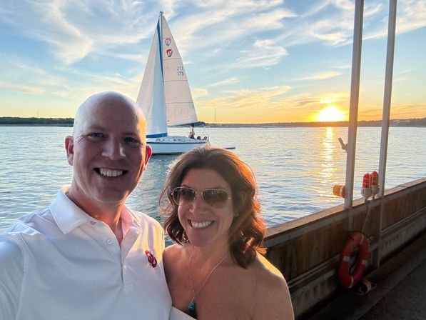 Smiling couple selfie on a wooden waterfront dock at sunset, sailboat passing on calm water with golden sun reflecting across the bay