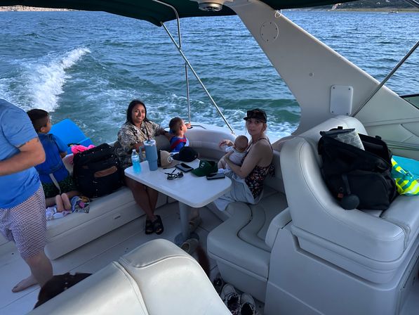Family on a motorboat cruising a lake, adults and children seated around a small cockpit table with drinks, bags, and the boat wake visible behind them.
