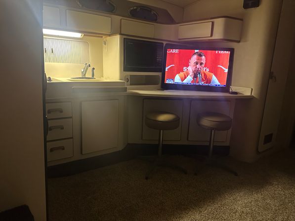 Cozy RV/motorhome kitchenette with sink, microwave, overhead cabinets, counter bar with two round swivel stools and a flat-screen TV showing a man speaking.