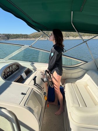 Person standing barefoot at the helm of a motorboat under a green bimini on a sunny lake, wearing sunglasses and a black jacket while steering with wooded shoreline and hills in the background.