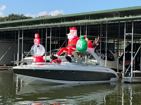 White-and-gray cabin boat tied in a covered marina slip, decorated for Christmas with a large inflatable Santa holding a green gift sack and a red snow-globe ornament on the bow, reflected in calm lake water.