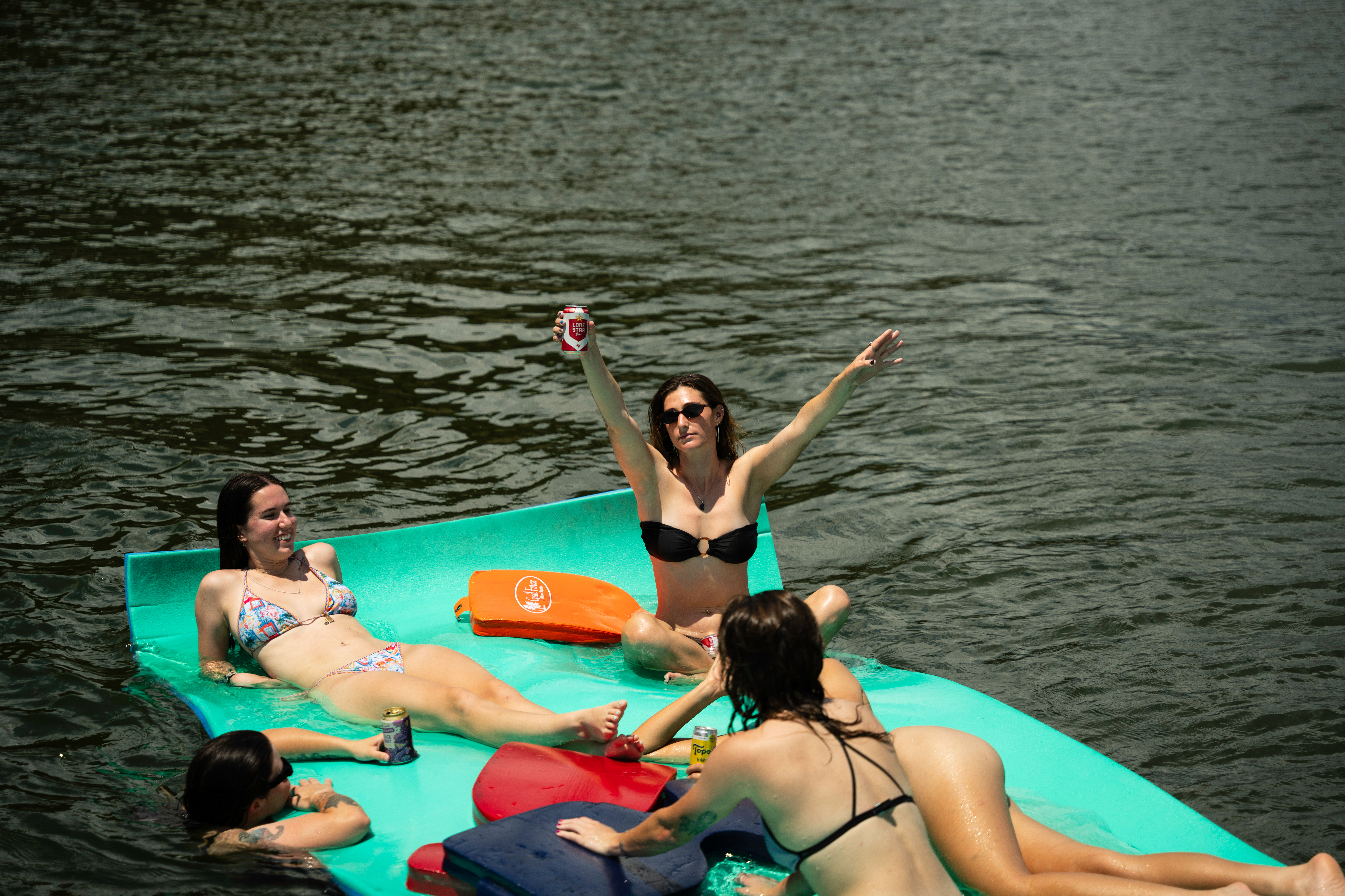 Group of women relaxing on a teal floating mat in a lake, wearing bikinis and holding canned drinks, one woman raising a can and cheering on a sunny summer day.