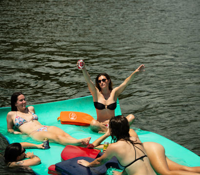 Group of women relaxing on a teal floating mat in a lake, wearing bikinis and holding canned drinks, one woman raising a can and cheering on a sunny summer day.