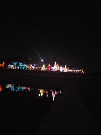 Festive waterfront holiday light display at night with illuminated trees, inflatable Santa and reindeer glowing and reflecting on calm water beside a dark dock.