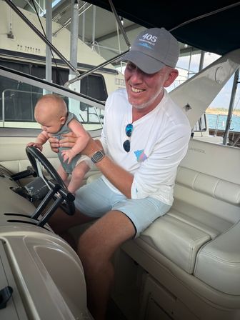Smiling man holding a baby at the helm of a docked motorboat in a sunny marina, baby reaching for the steering wheel.