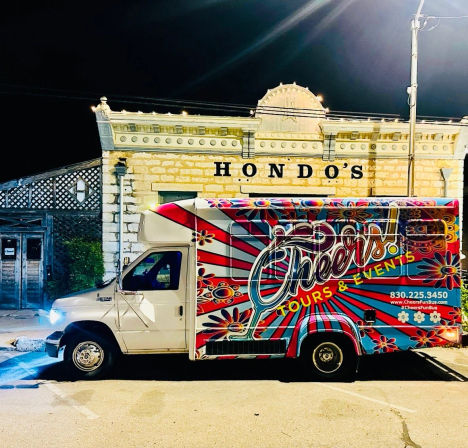 Vibrant floral-painted party bus parked at night on a downtown main street in front of an illuminated historic stone storefront with decorative cornice