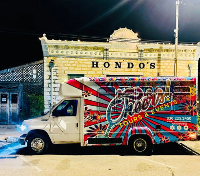 Vibrant floral-painted party bus parked at night on a downtown main street in front of an illuminated historic stone storefront with decorative cornice