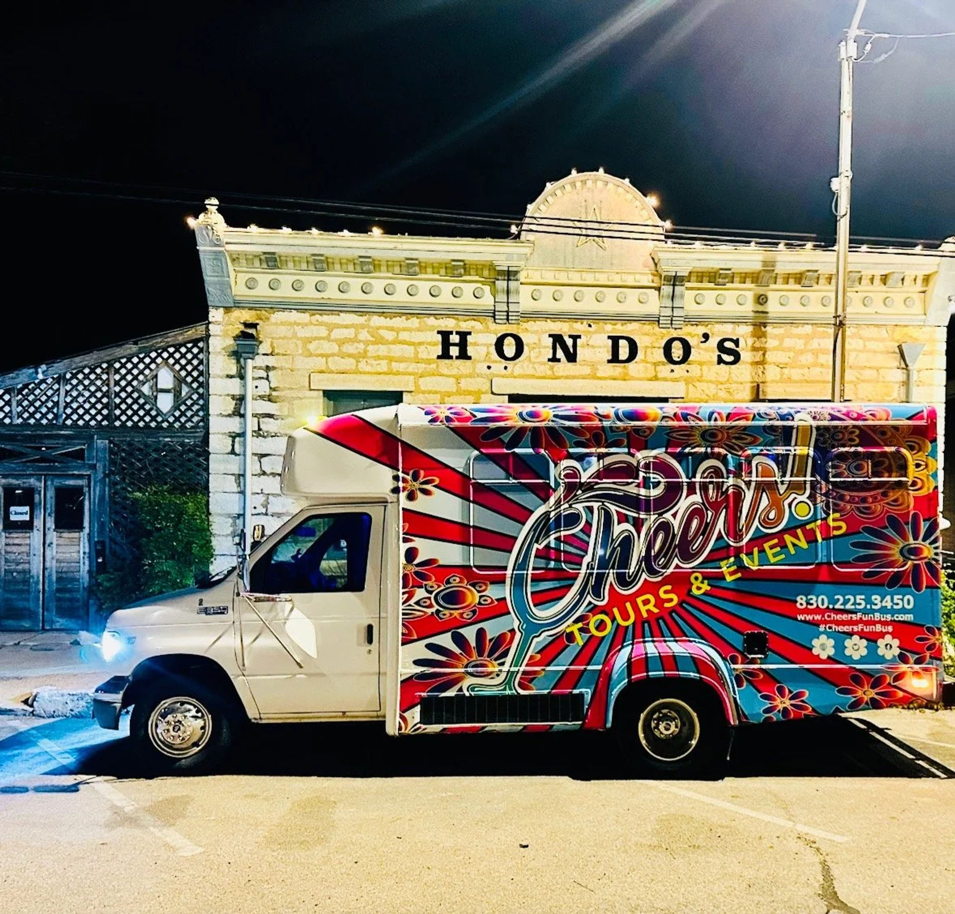 Vibrant floral-painted party bus parked at night on a downtown main street in front of an illuminated historic stone storefront with decorative cornice