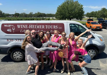 Cheerful group of women in pink outfits lifting a laughing man in front of a white wine-tour shuttle van parked by vineyard rows on a sunny day
