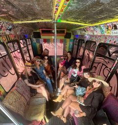 Group of women in hats and sunglasses enjoying a boho-style party bus interior with patterned sofas, a center dance pole, sticker-covered walls and colorful bunting.