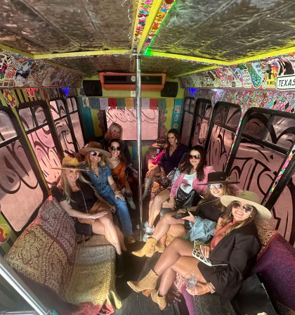 Group of women in hats and sunglasses enjoying a boho-style party bus interior with patterned sofas, a center dance pole, sticker-covered walls and colorful bunting.