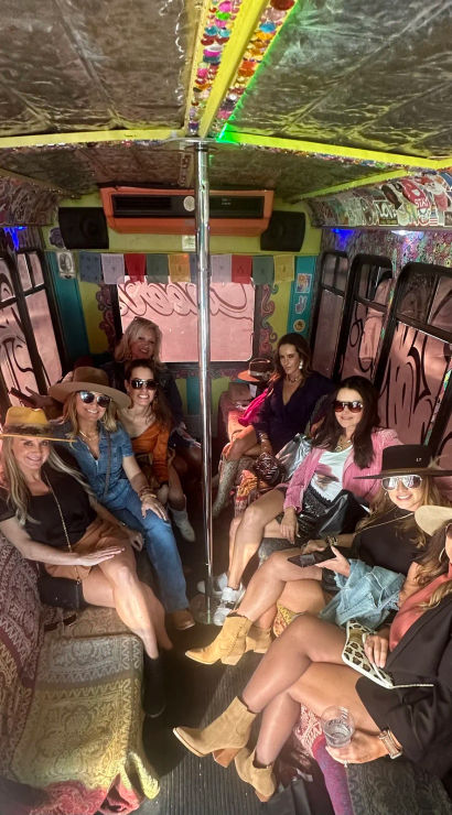 Group of women in hats and sunglasses enjoying a boho-style party bus interior with patterned sofas, a center dance pole, sticker-covered walls and colorful bunting.