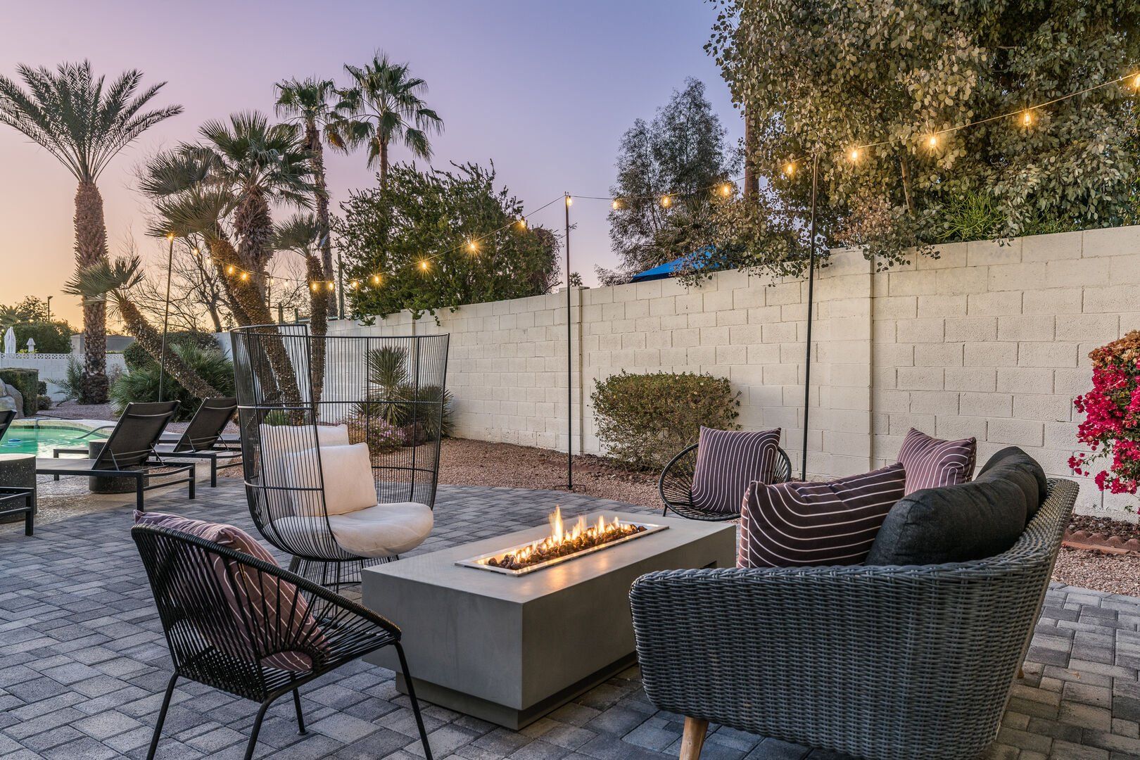 Cozy modern backyard patio at dusk with wicker seating and striped pillows around a rectangular gas fire pit, glowing string lights overhead, palm trees and a pool beside a cinderblock privacy wall.