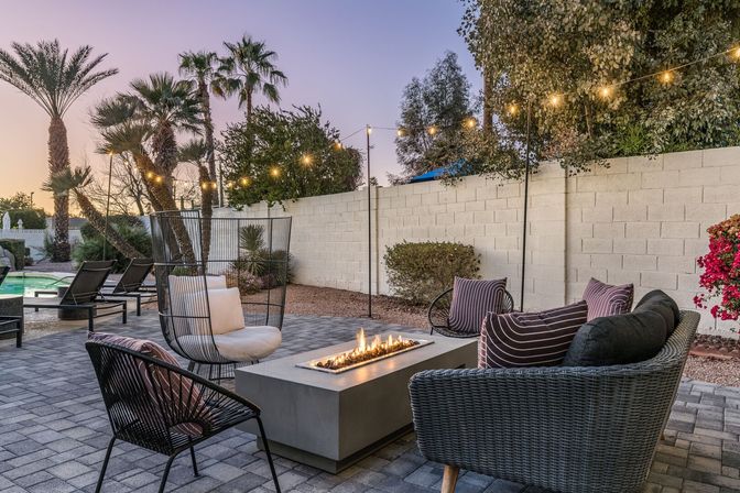 Cozy modern backyard patio at dusk with wicker seating and striped pillows around a rectangular gas fire pit, glowing string lights overhead, palm trees and a pool beside a cinderblock privacy wall.