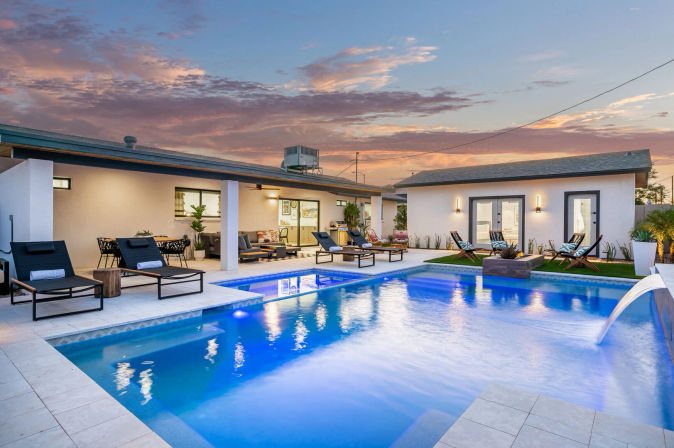 Resort-style modern backyard oasis at dusk with an illuminated blue swimming pool, cascading water feature, sun loungers, covered patio seating and dining under a colorful sunset sky.