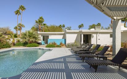 Sun-soaked backyard desert oasis with a swimming pool, row of wicker sun loungers under a slatted pergola, palm trees and a white mid-century modern home.