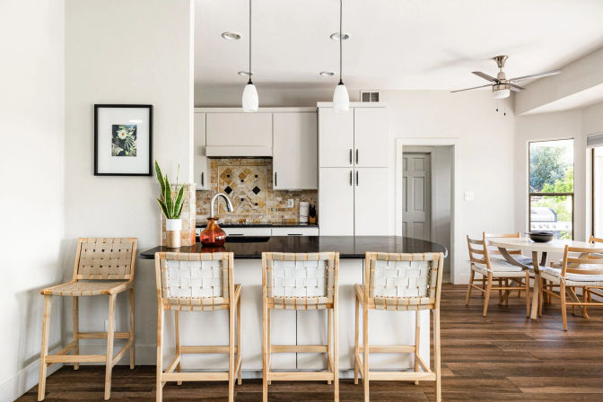 Bright modern open-plan kitchen with dark countertop island and breakfast bar, four woven wood bar stools, white cabinets, mosaic tile backsplash, pendant lights, wood floors and adjacent dining nook with outdoor view