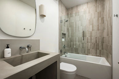 Minimalist modern bathroom in neutral tones — concrete-style sink with wall-mounted chrome faucet, oval mirror, white toilet, and glass-enclosed bathtub-shower with beige vertical tiles