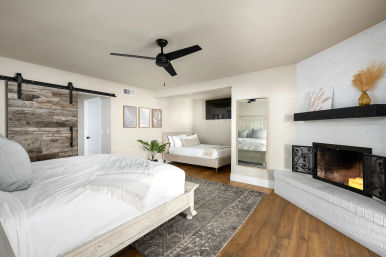 Bright modern master bedroom with white linens on a wooden bed, rustic sliding barn door, black ceiling fan, full-length mirror, potted plant, and white brick fireplace with candles on hardwood floors.