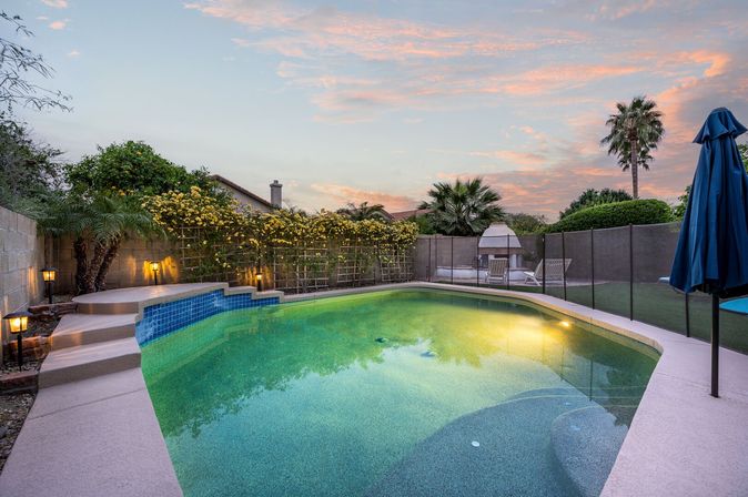 Backyard swimming pool at sunset with glowing water, tiled steps, safety fence, yellow flowering vine, palm trees, lounge chairs and an outdoor fireplace