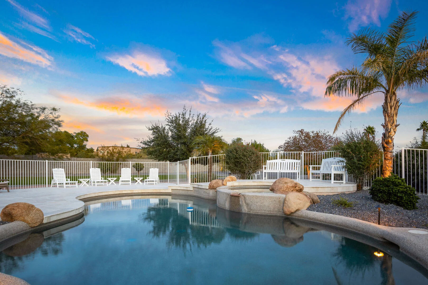 Backyard swimming pool and spa at sunset with a palm tree, white lounge chairs, rock accents, and a white safety fence.