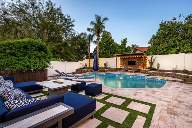 Modern backyard outdoor living space with blue cushioned lounge seating, geometric stone-and-grass pavers, a tiled swimming pool, palm trees and a lit cabana with chaise lounges at dusk.
