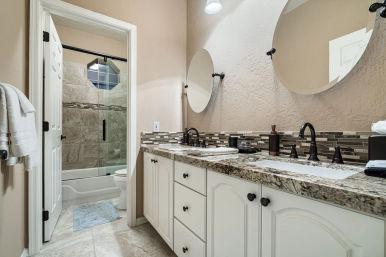Neutral beige residential bathroom with granite double-sink vanity, white cabinets, round wall mirrors, dark oil-rubbed faucets, mosaic tile backsplash, and glass-enclosed tiled tub/shower with octagonal window.