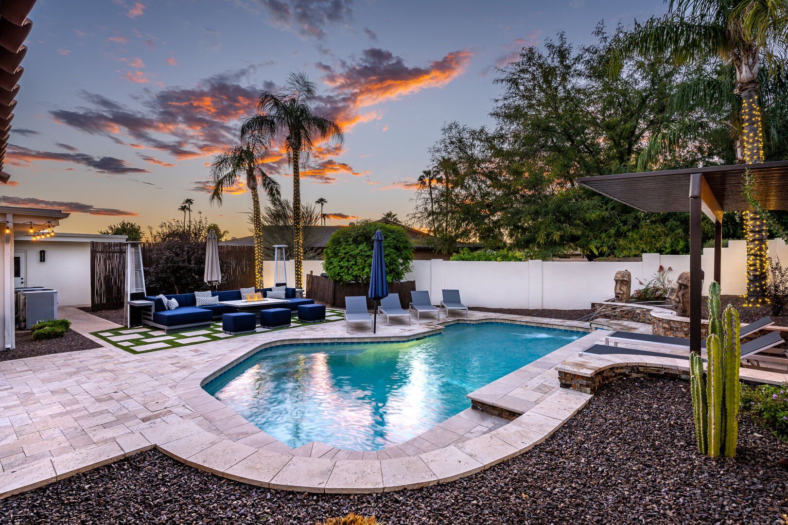Twilight backyard pool scene with turquoise swimming pool, stone tile patio, navy outdoor sectional and loungers, palm trees wrapped in string lights, cactus and pergola under a colorful sunset sky.