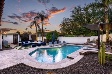 Twilight backyard pool scene with turquoise swimming pool, stone tile patio, navy outdoor sectional and loungers, palm trees wrapped in string lights, cactus and pergola under a colorful sunset sky.