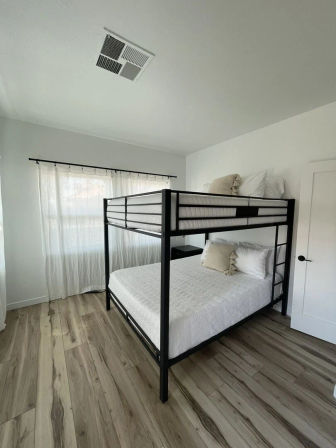 Bright minimalist bedroom with black metal bunk bed (queen bottom, twin top), crisp white linens, sheer curtains, and light wood-look flooring
