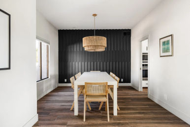 Modern dining room with a white rectangular table, six cane-back wooden chairs, woven rattan pendant light, textured black accent wall and dark wood plank flooring.