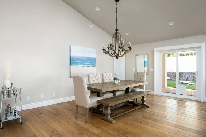 Bright coastal-style dining room with rustic wooden farmhouse table and bench, tufted beige chairs, chandelier, hardwood floors, ocean artwork, and sliding glass doors to a sunny backyard.