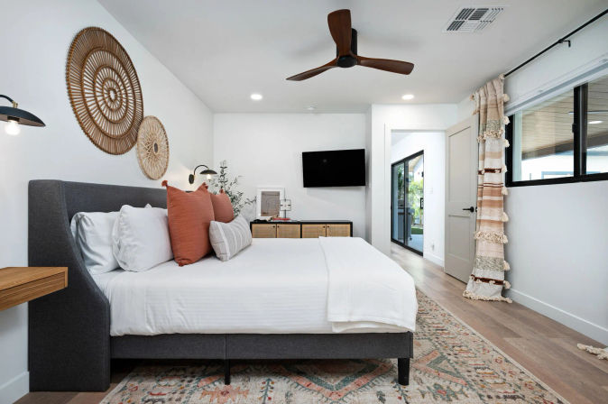 Bright modern bedroom interior with a gray upholstered king bed, white linens and rust throw pillows, woven boho wall decor, wooden ceiling fan, wall-mounted TV, rattan dresser and sliding glass doors to an outdoor patio.