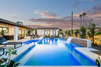 Modern backyard swimming pool at sunset with a cascading waterfall feature, lounge chairs, patio seating and palm trees.