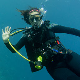Scuba diver waving underwater in clear blue ocean, wearing mask, regulator, black wetsuit and buoyancy compensator with a bright yellow hose