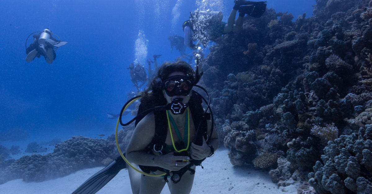 Scuba diver in full gear hovering above a sandy seabed beside a coral reef, bubbles rising as a small group of divers explores clear blue tropical ocean water.