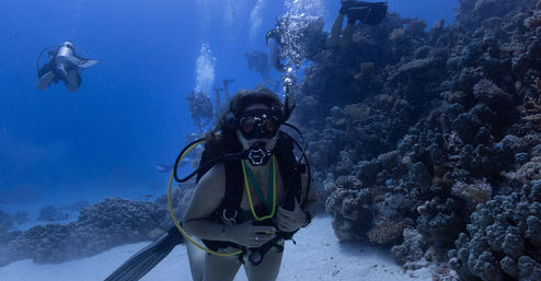 Scuba diver in full gear hovering above a sandy seabed beside a coral reef, bubbles rising as a small group of divers explores clear blue tropical ocean water.