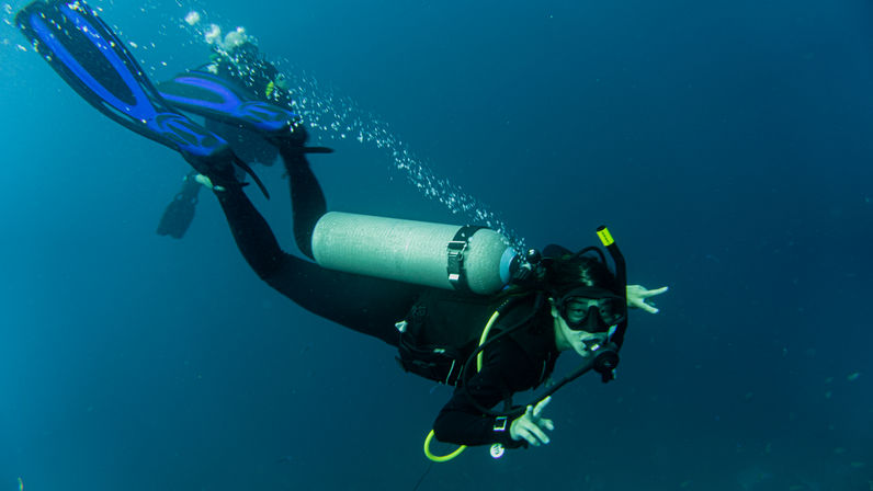 Scuba diver gliding through deep blue ocean with silver air tank and blue fins, bubbles trailing as they flash an OK hand signal