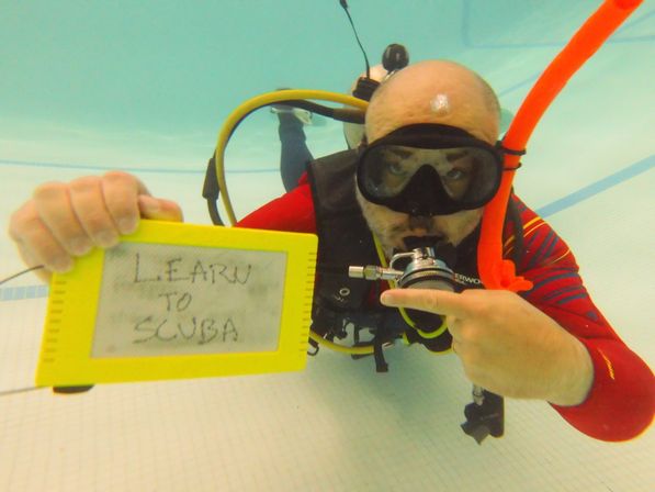 Underwater scuba training in an indoor pool: diver in mask and regulator points to a yellow slate reading 'LEARN TO SCUBA'.