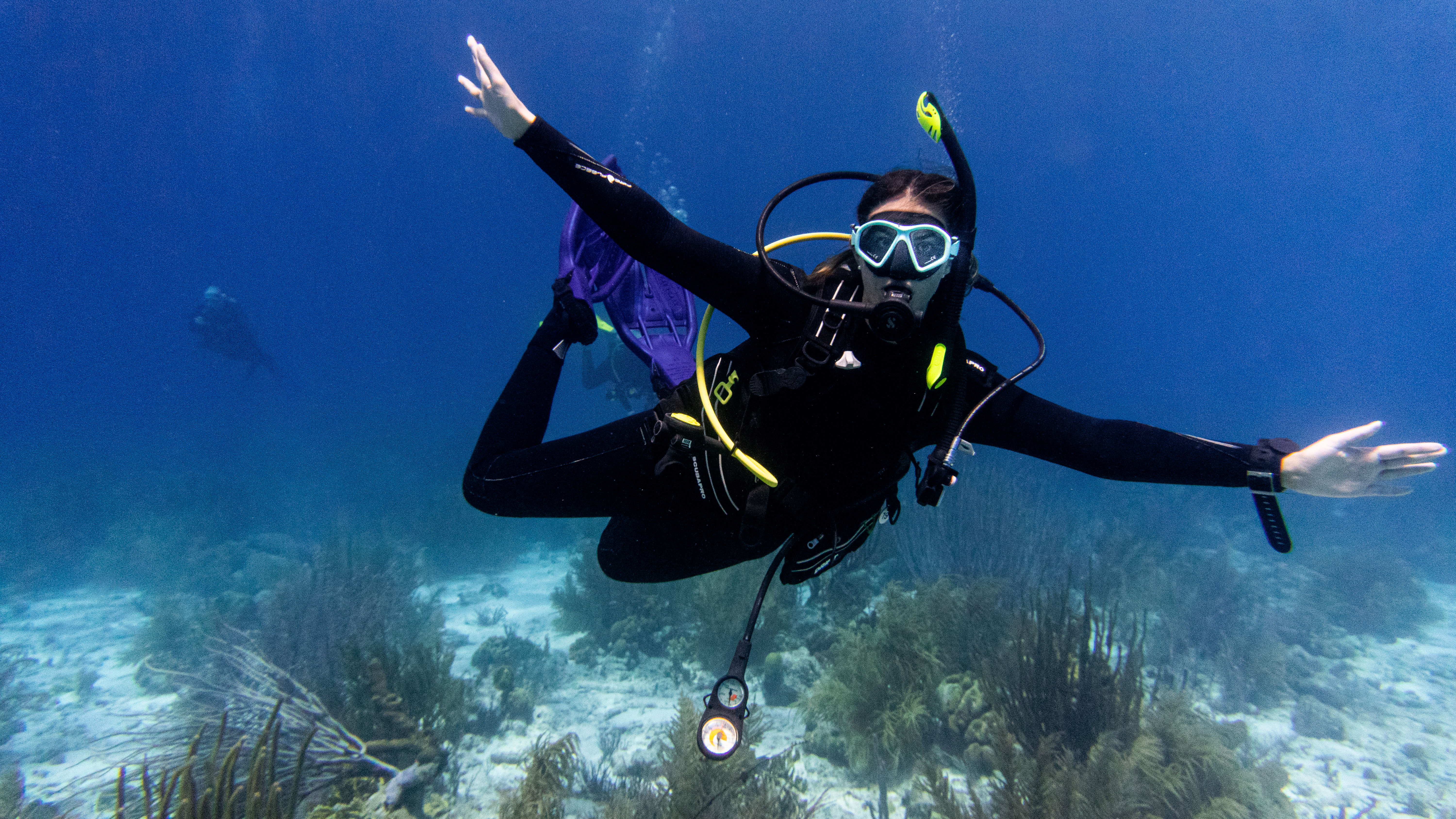Scuba diver in black wetsuit and purple fins gliding with arms outstretched over a shallow tropical coral reef in clear blue ocean water, wearing mask, regulator, and dive gauges
