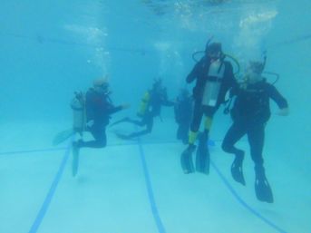 Group of scuba divers in tanks, masks, and fins practicing underwater in a clear indoor swimming pool during a dive training session