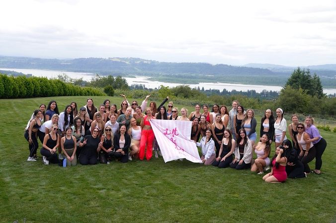 Large group of women smiling and posing on a grassy hillside holding a pink "Lift" banner at an outdoor fitness/wellness retreat with a river and rolling hills in the background.