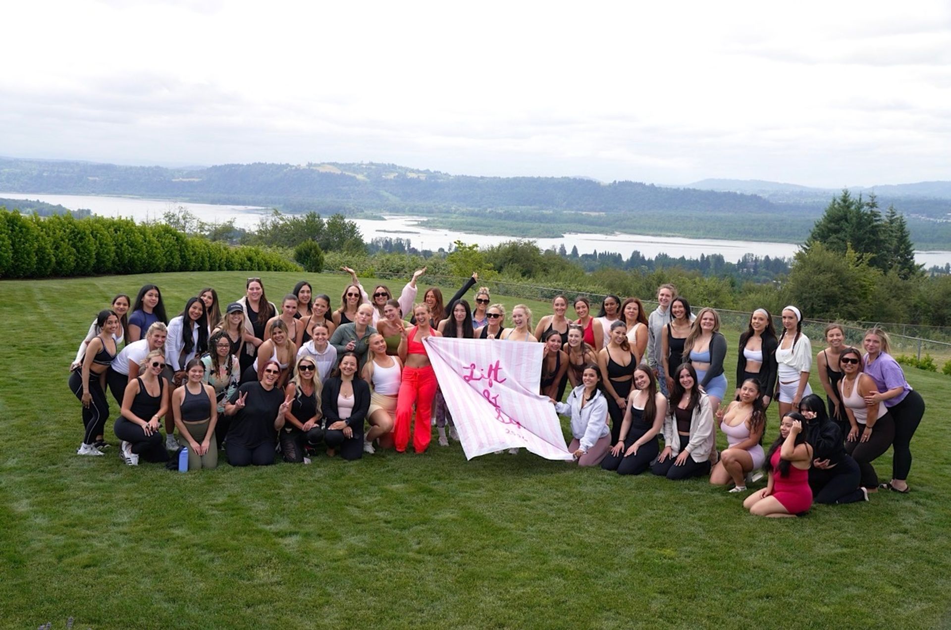Large group of women smiling and posing on a grassy hillside holding a pink "Lift" banner at an outdoor fitness/wellness retreat with a river and rolling hills in the background.