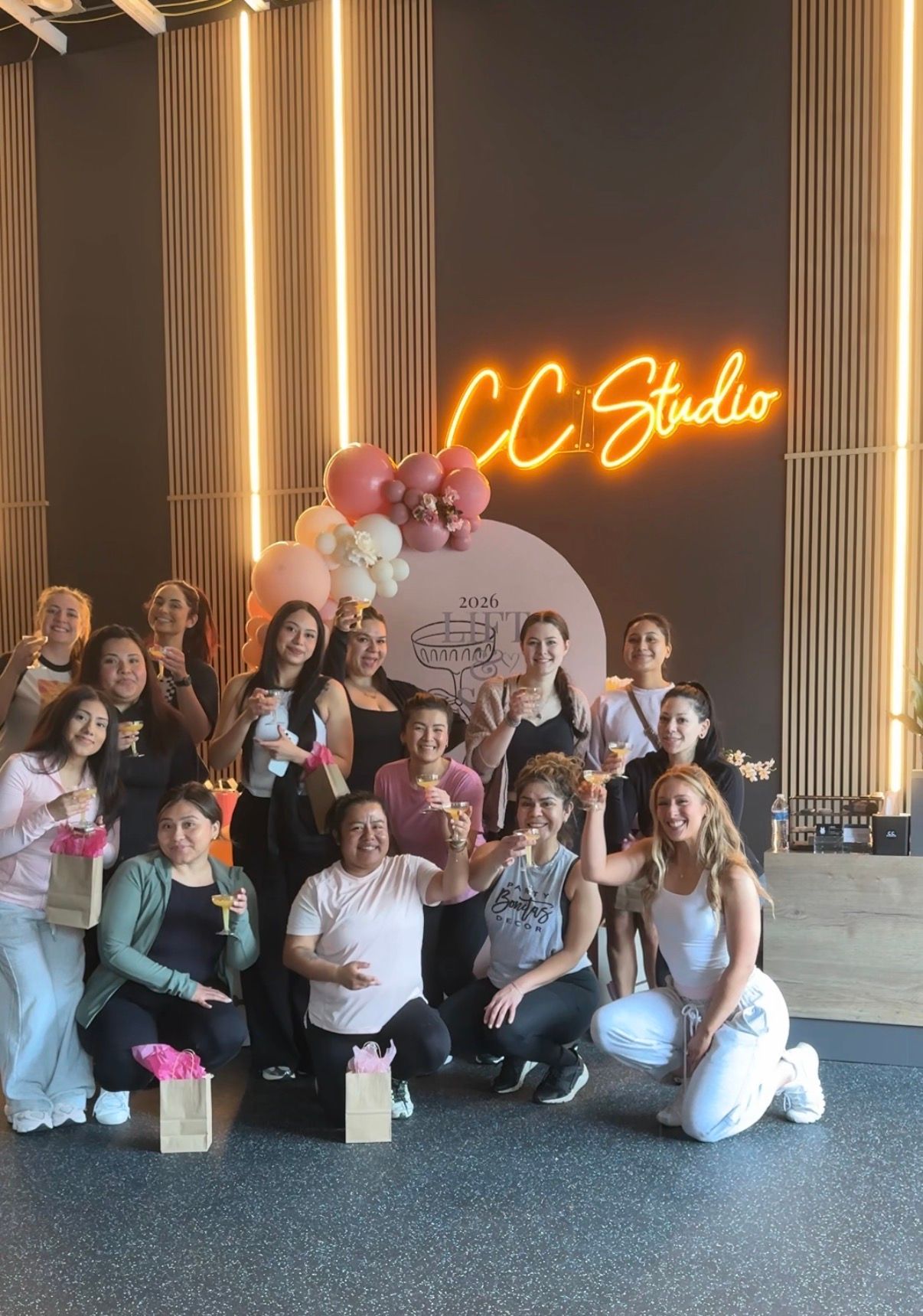 Group of women celebrating inside a modern studio with a pink balloon arch, gift bags, and raised champagne glasses beneath a glowing neon wall sign.